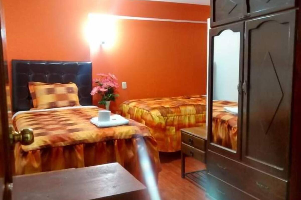 Une chambre avec deux lits, un banc en bois, une armoire et une lampe fixée au mur.