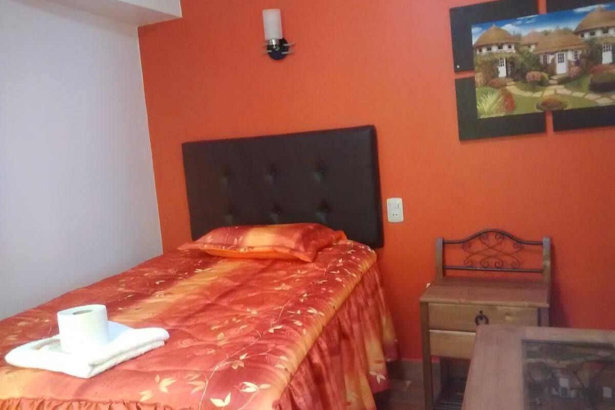 Une chambre à coucher avec un lit, une table de chevet, une lampe fixée au mur et un tableau encadré.