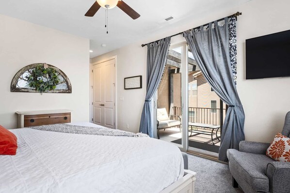 A bedroom with a bed, a nightstand, a ceiling fan, a window with a wreath, and a sliding glass door leading to an outdoor area with a chair and a table.