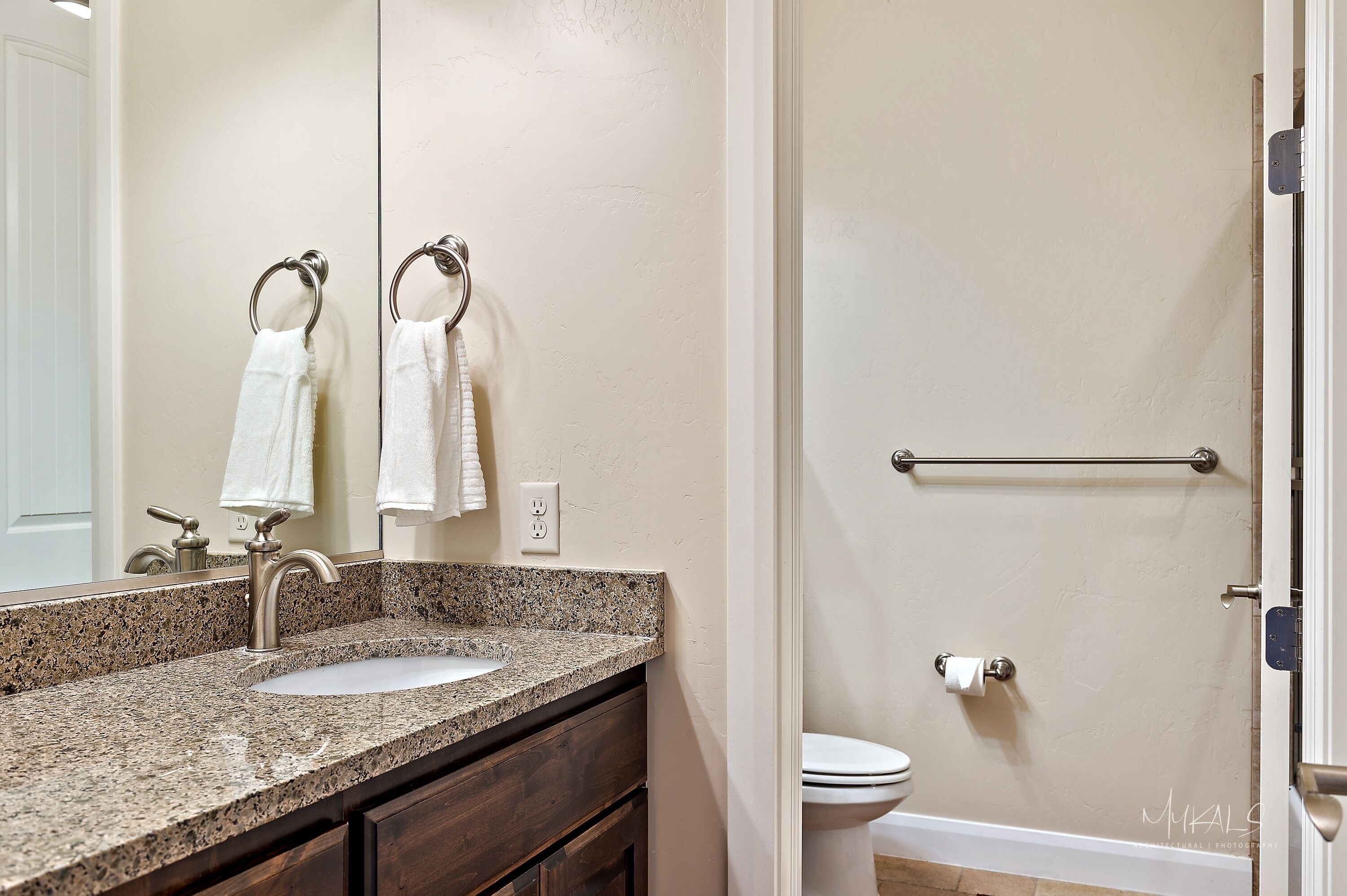 Une salle de bain avec un lavabo doté d’un plan de travail en granit, un miroir, deux serviettes suspendues à des porte-serviettes, des toilettes et une barre d’appui près des toilettes.