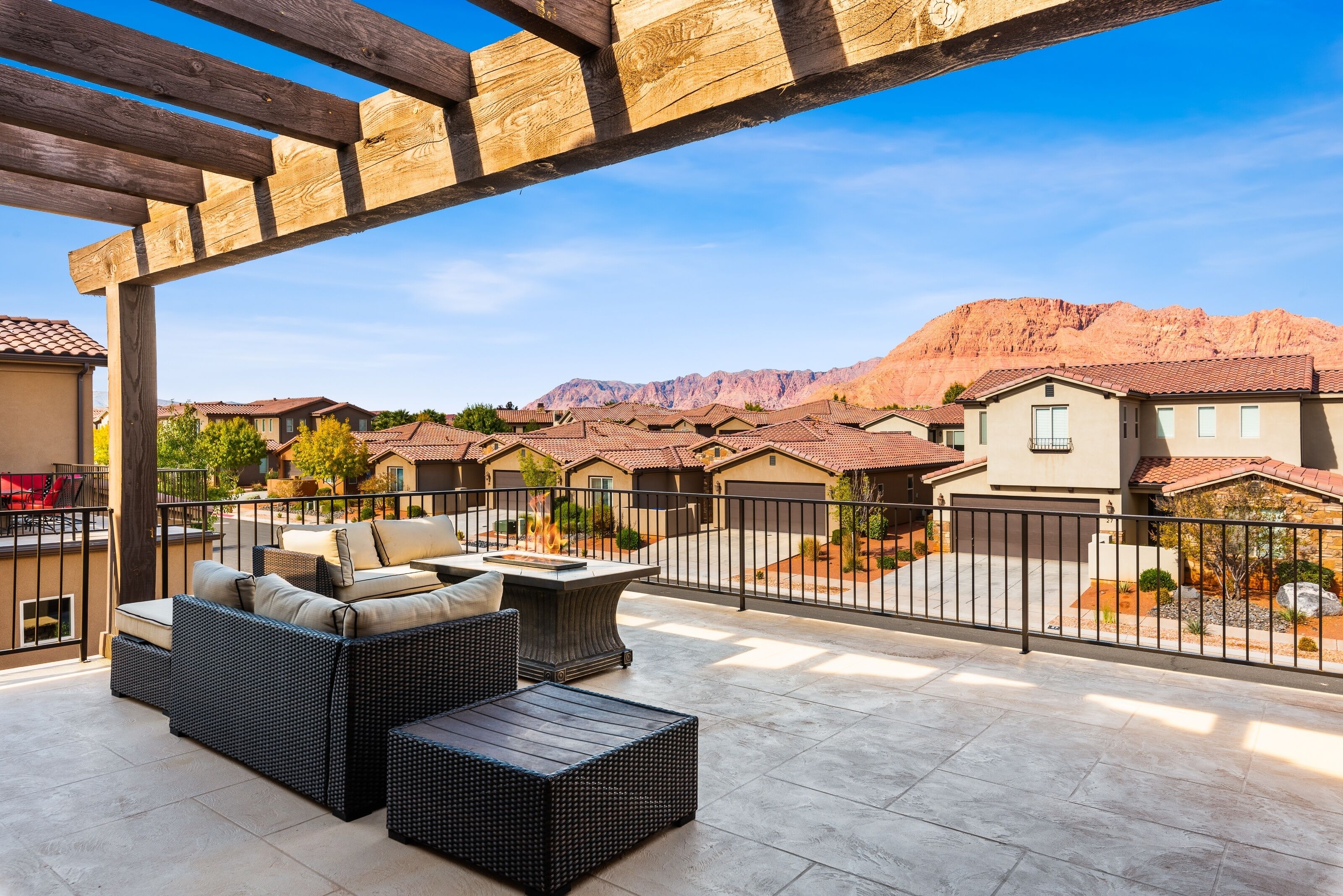 A covered patio with wicker furniture, a fire pit, and a view of a residential area with houses and mountains.