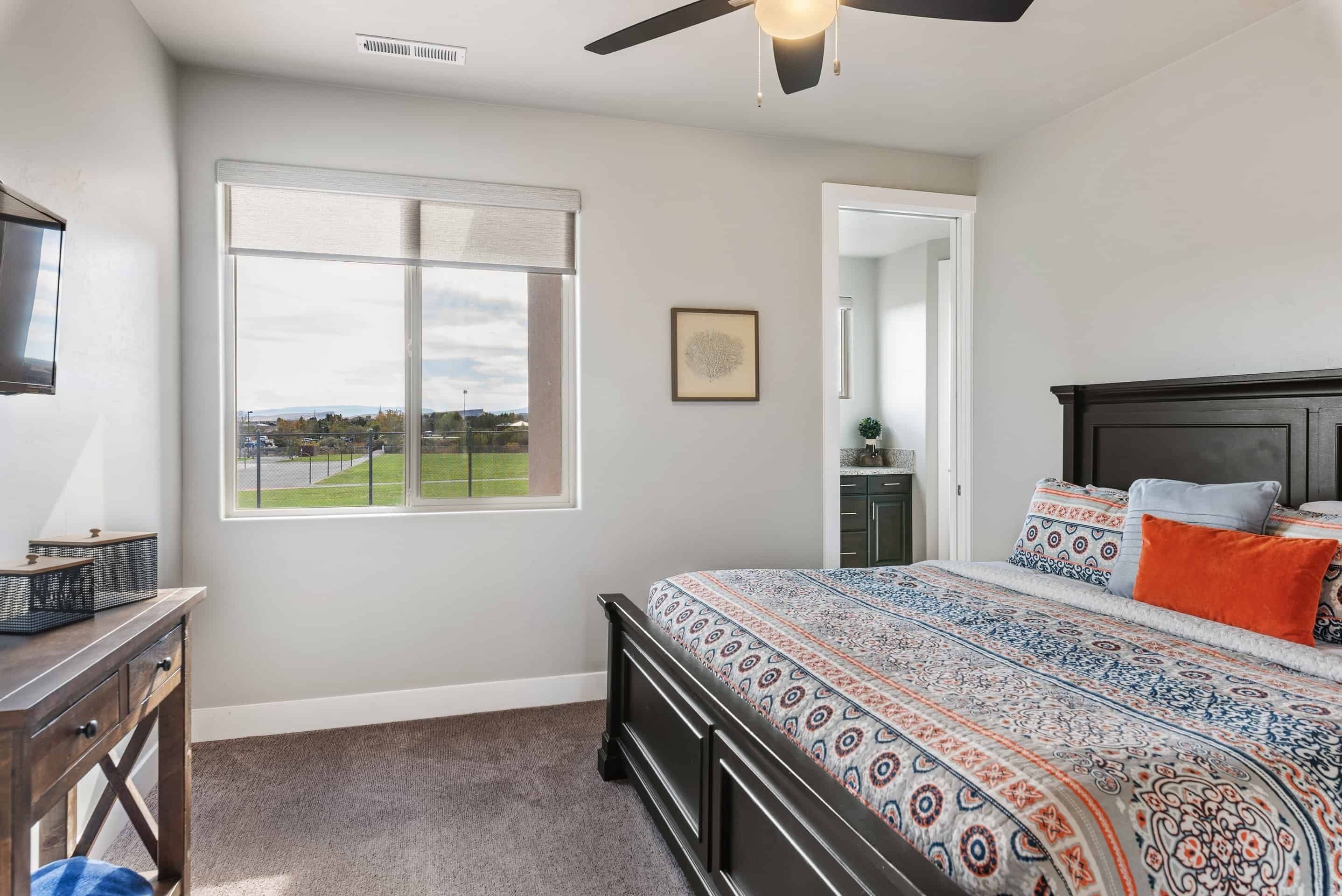 A bedroom with a large window, a bed with patterned bedding, a ceiling fan, and a view of a park.