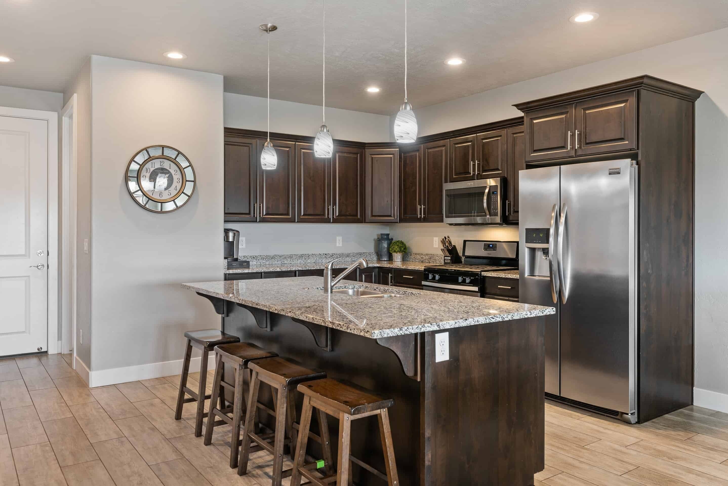 A modern kitchen with dark wood cabinets, stainless steel appliances, a granite countertop island with bar stools, pendant lighting, and a wall clock.