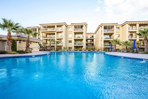 A swimming pool with a clear blue water surface, surrounded by a multi-story building with balconies and palm trees.