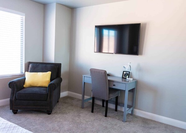 A room with a black armchair with a yellow pillow, a grey desk with a chair, and a flat-screen TV mounted on the wall.
