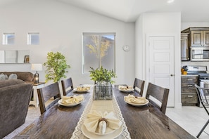 Enjoy meals in this stylish dining area with a long table, sofa, and side table, offering a view of a tree through the large window.