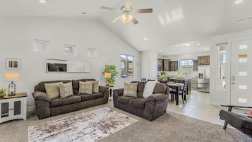 A spacious living room with a high ceiling, a ceiling fan, and a dining area with a kitchen in the background.