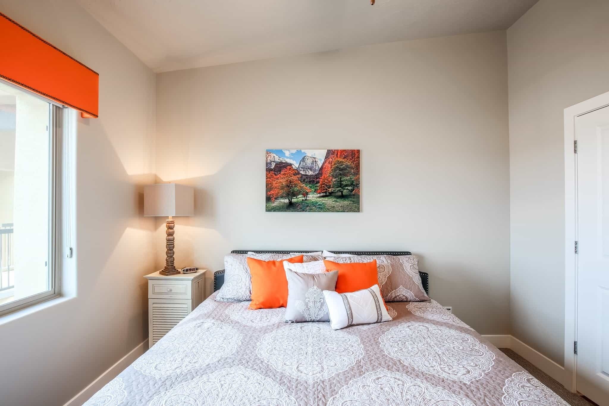 A bedroom with a bed, pillows, a nightstand, a lamp, a window, and a painting on the wall.
