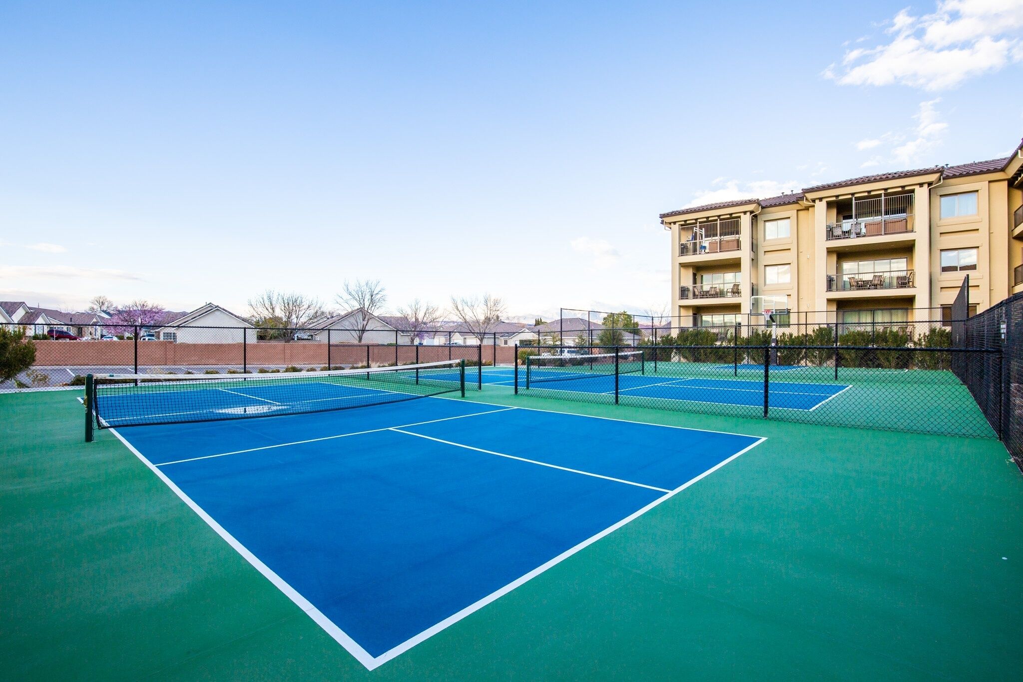 A tennis court with a net, surrounded by a fence, and a building with balconies in the background.