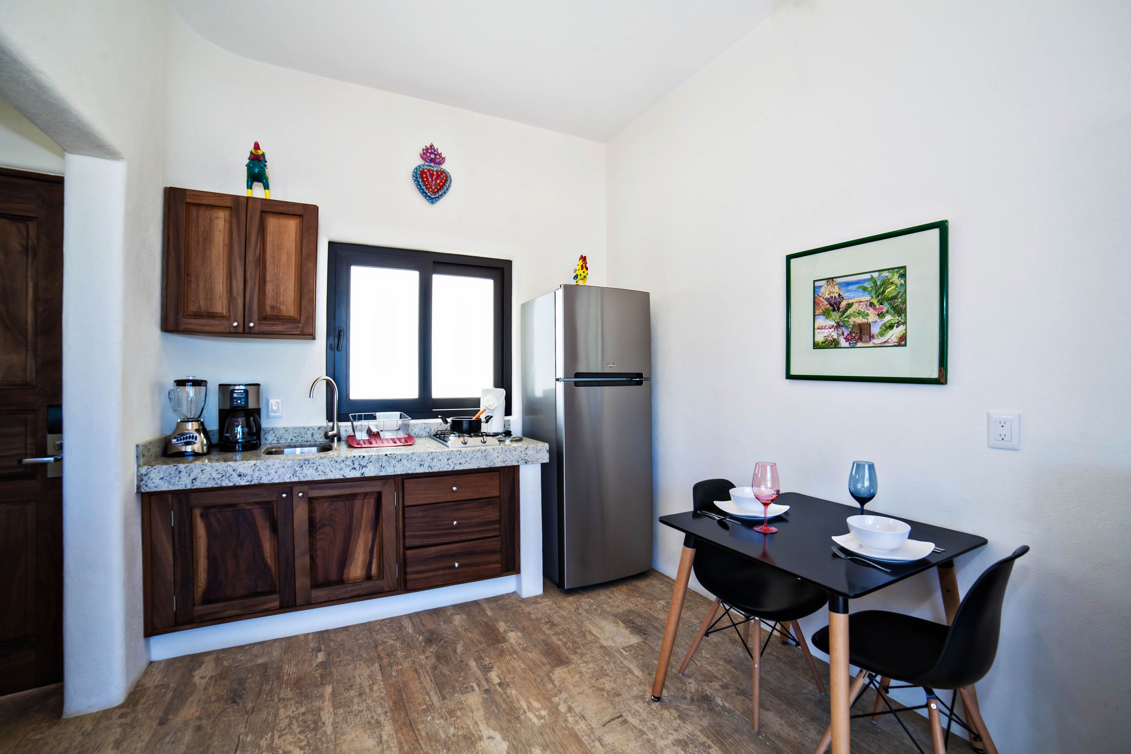 A modern kitchen with dark wood cabinets, a stainless steel refrigerator, a granite countertop, and a dining area with a black table and chairs.