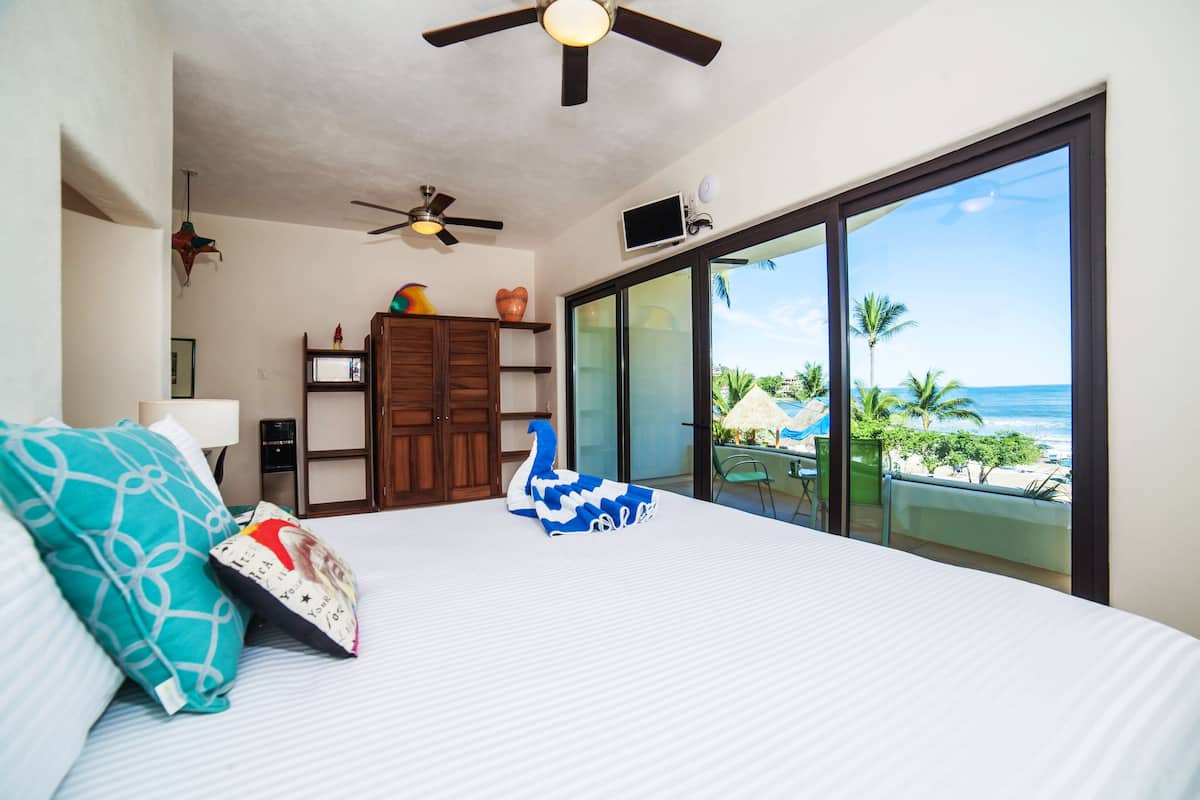 A bedroom with a large bed, a ceiling fan, a wooden cabinet, and a view of the beach through large windows.
