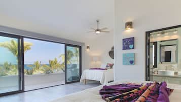 Un dormitorio espacioso con una cama grande, ventilador de techo y vistas a palmeras y al océano a través de ventanas de pared a techo.