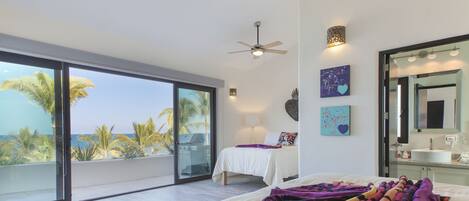 Un dormitorio espacioso con una cama grande, ventilador de techo y vistas a palmeras y al océano a través de ventanas de pared a techo.