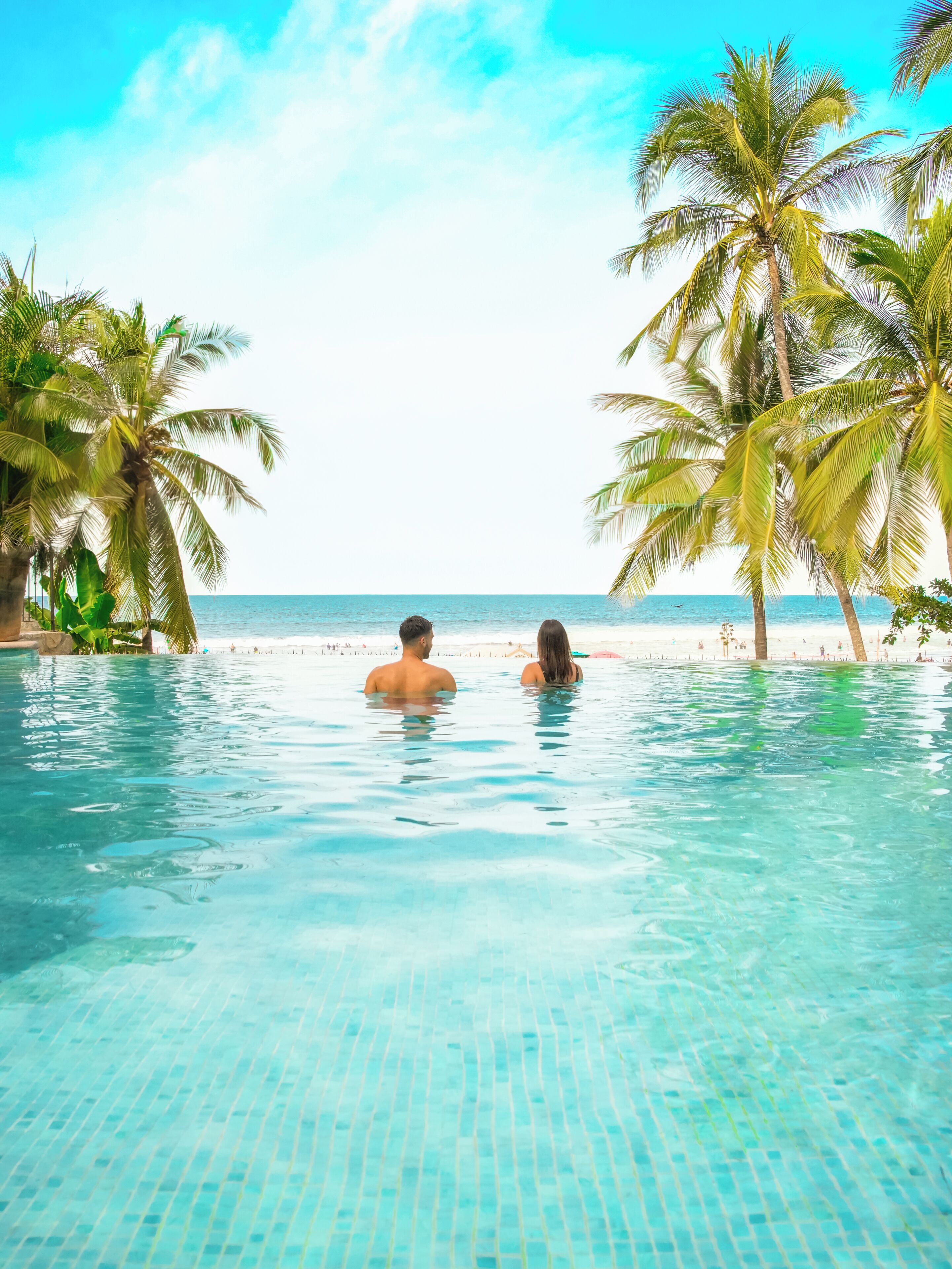 Duidelijk uitzicht op een zwembad met twee personen in het water, omringd door palmbomen en een strand in de verte.