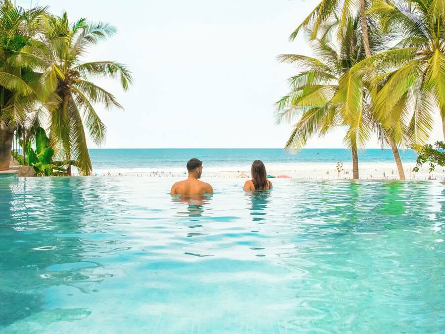 On voit clairement une piscine avec deux personnes dans l’eau, entourée de palmiers et avec une plage en arrière-plan.