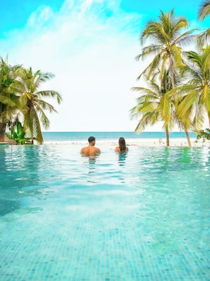 Vista despejada de una piscina con dos personas nadando, rodeada de palmeras y una playa al fondo.