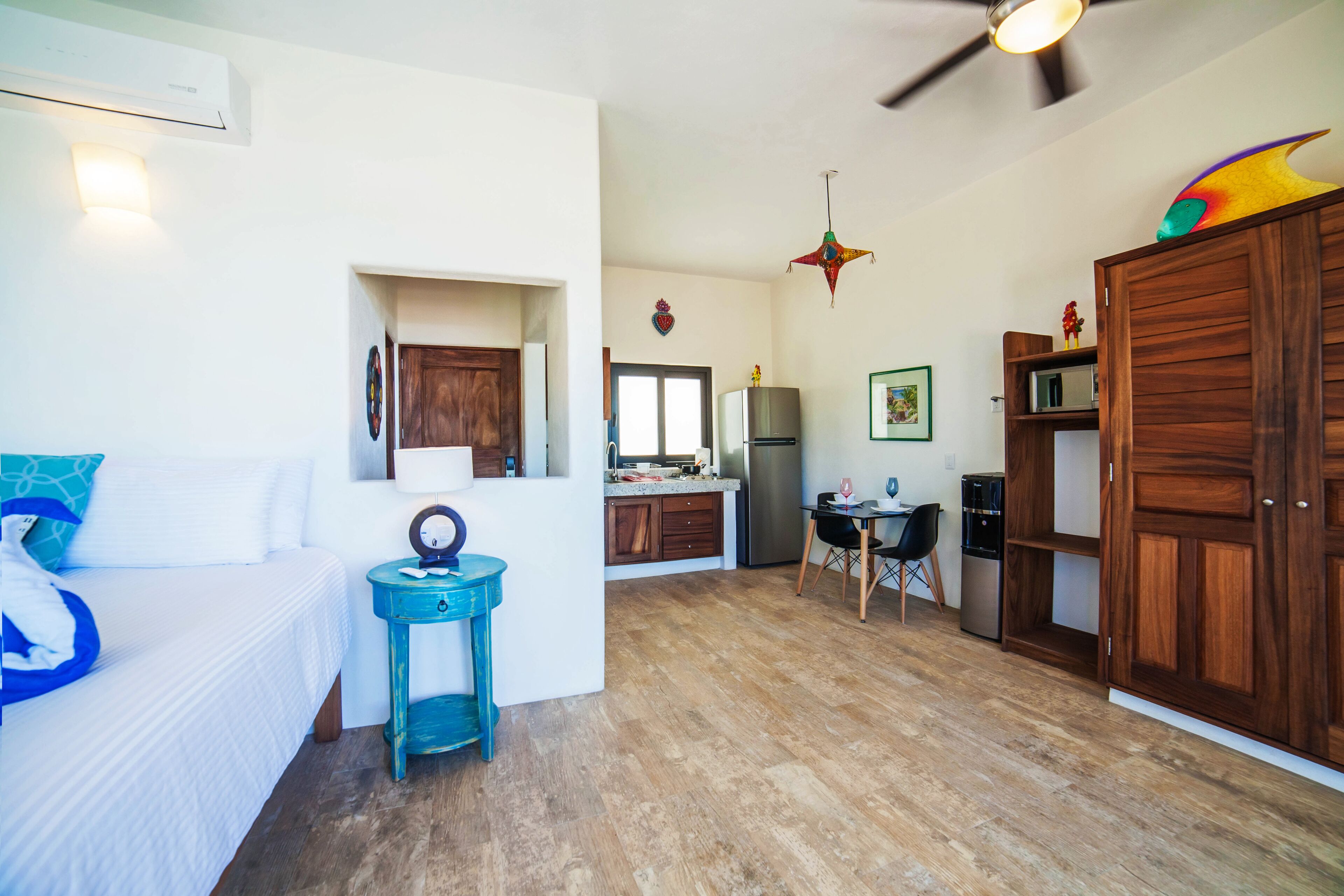 A spacious room with a bed, a small table, a ceiling fan, and a kitchen area with a refrigerator and cabinets.