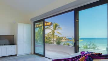 Een kamer met een grote glazen schuindeur naar een balkon met uitzicht op een strand met palmbomen en de oceaan.