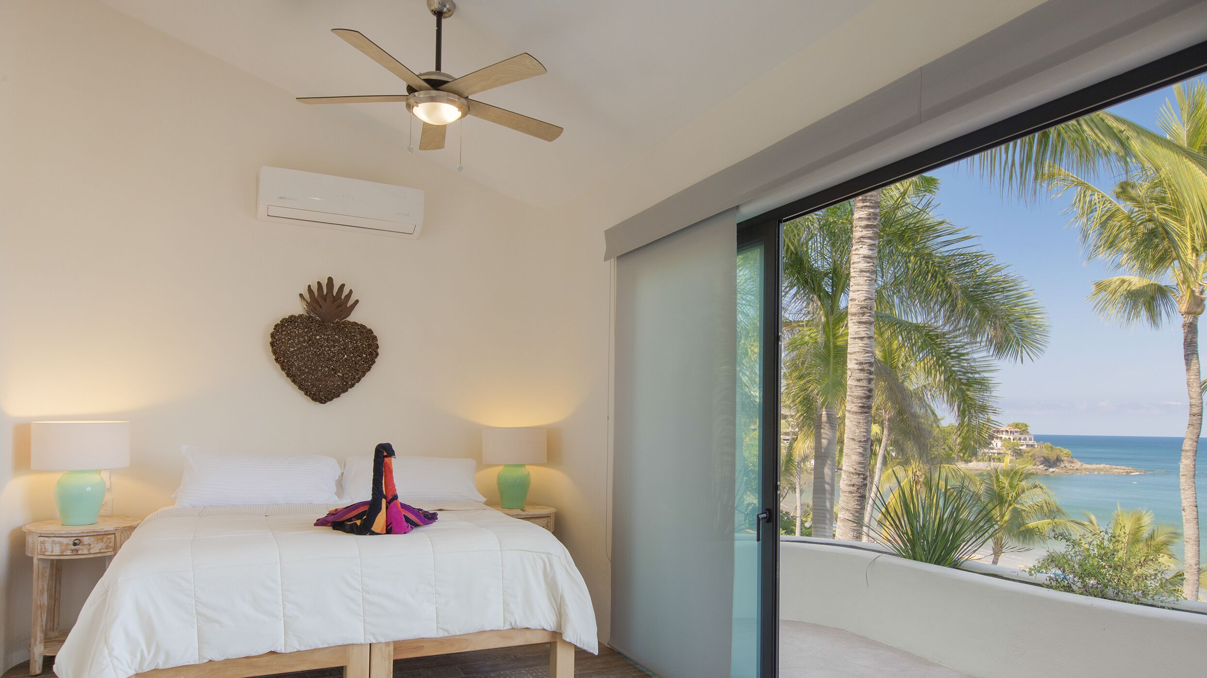 Un dormitorio con cama, ventilador de techo, decoración en forma de corazón en la pared y vista a palmeras y al océano.