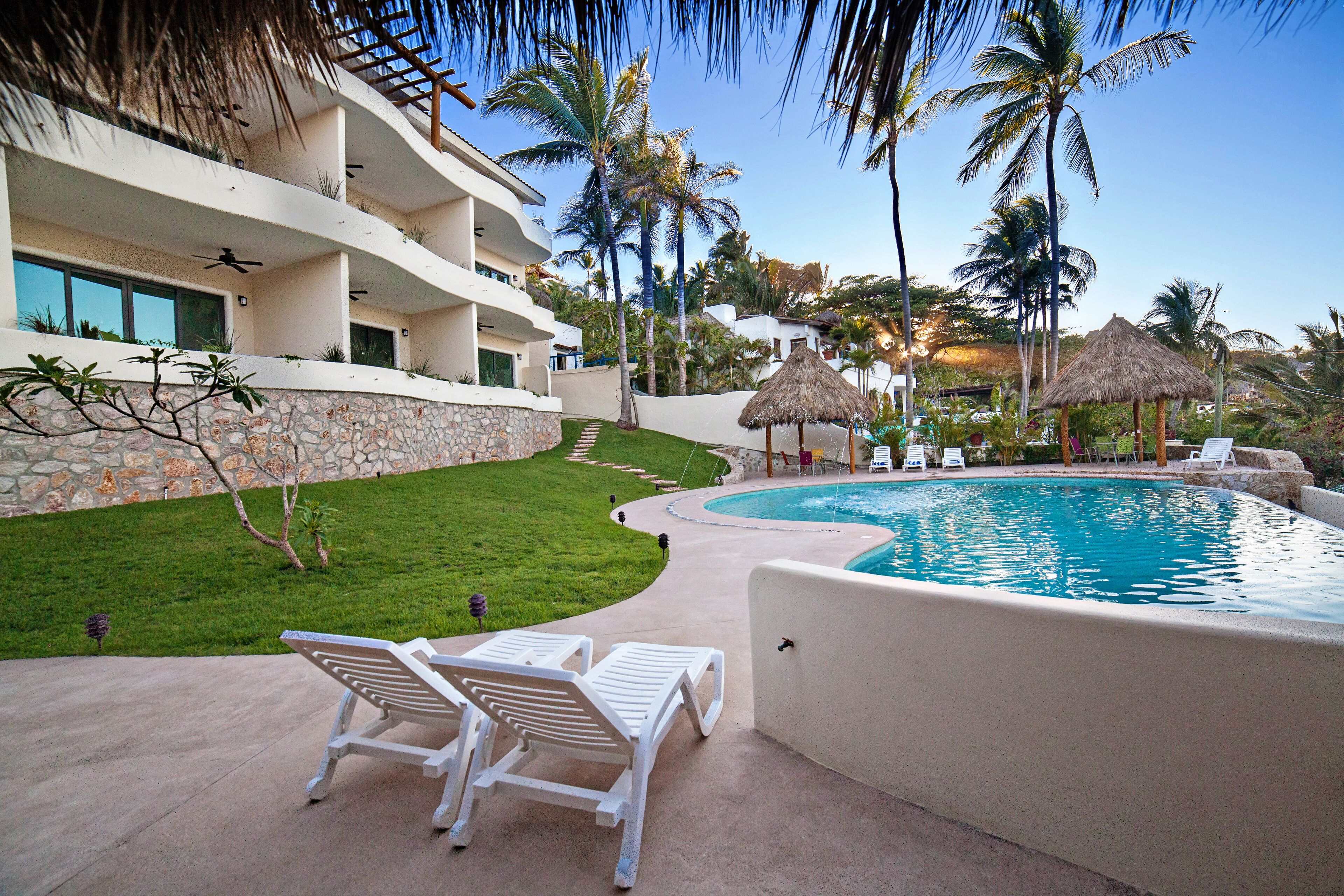 A resort with a swimming pool, white lounge chairs, palm trees, and a stone wall.