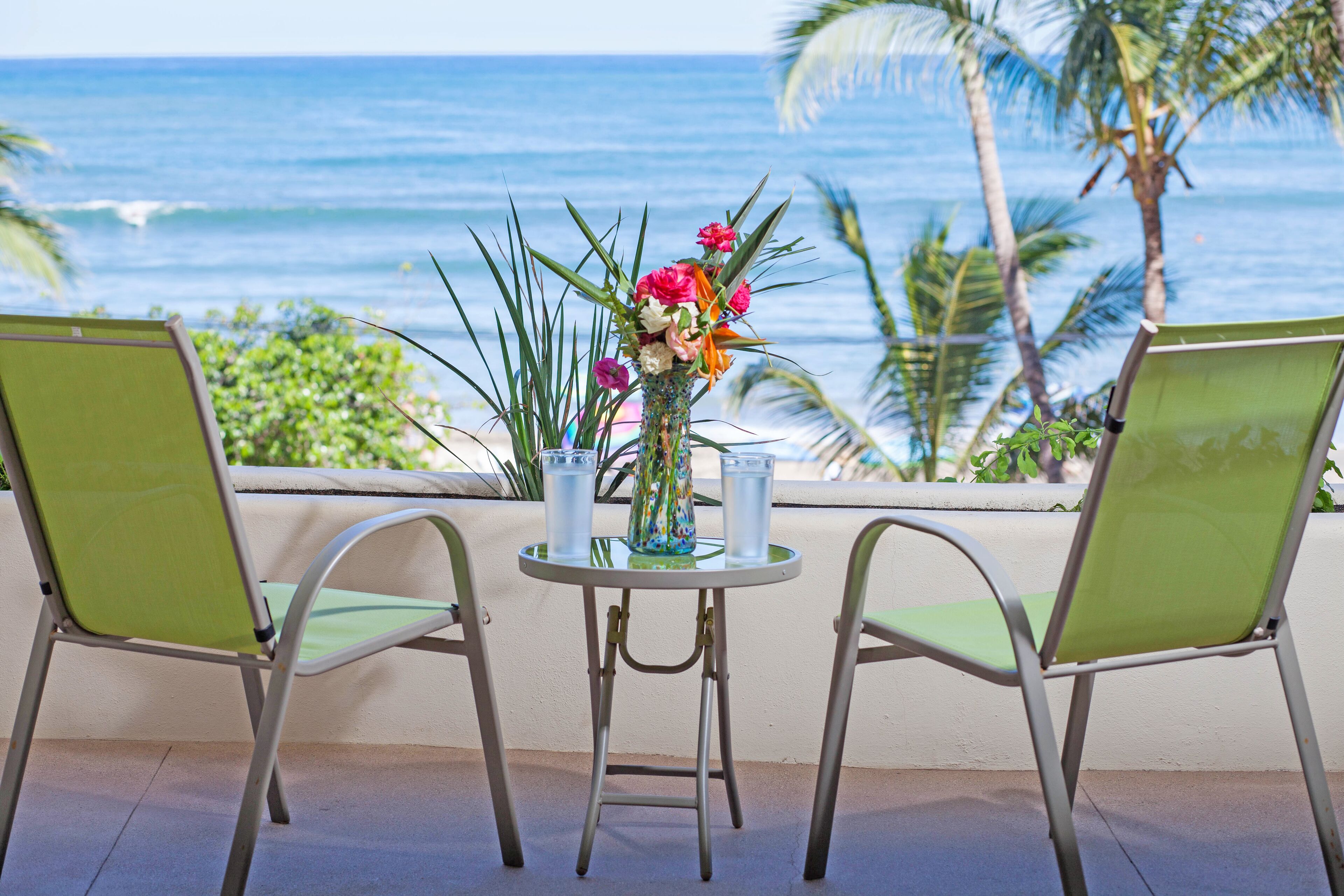 A table with two chairs and a vase of flowers, overlooking a beach with palm trees and the ocean.