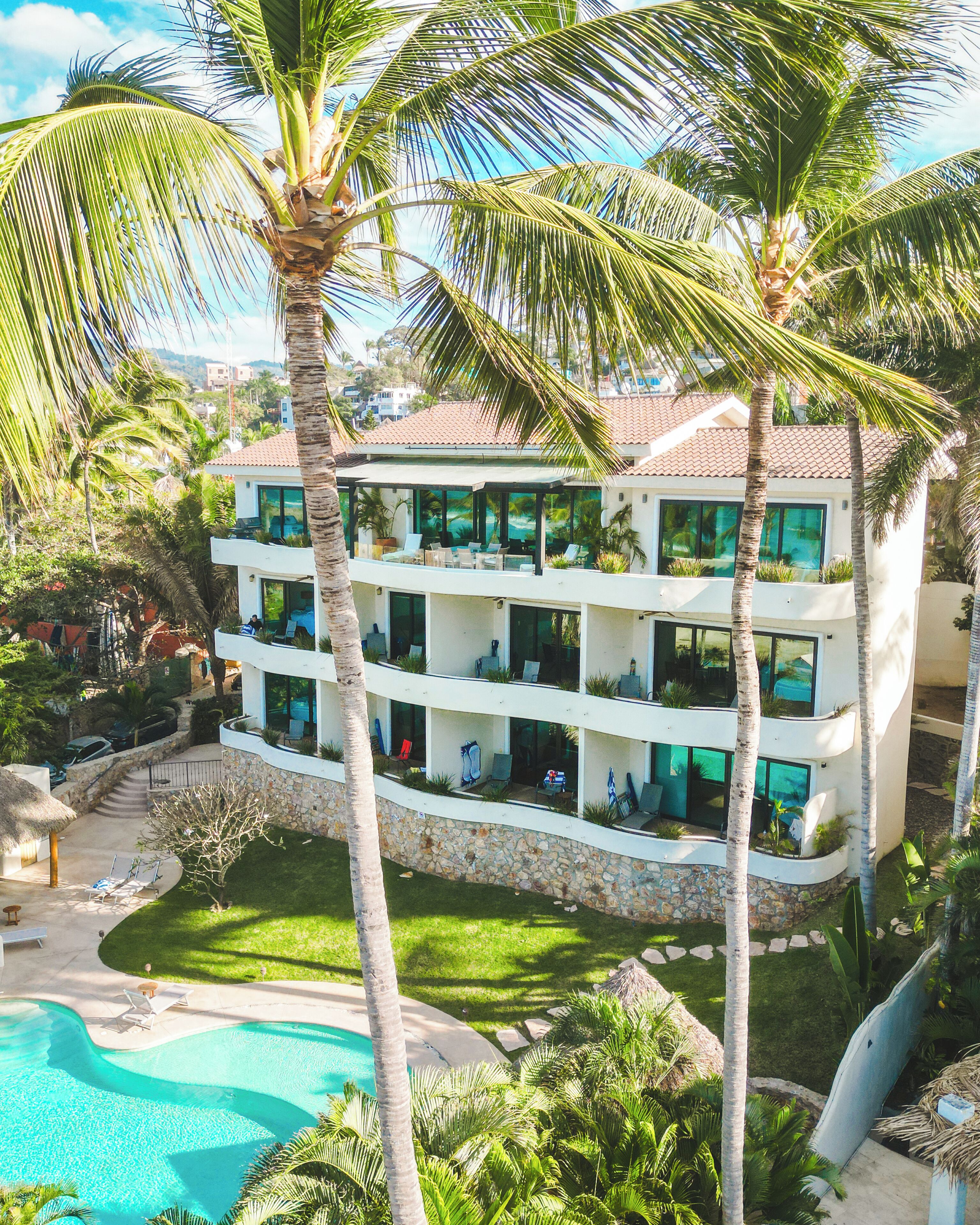 Un hotel de varios pisos con balcones, piscina y exuberante vegetación tropical.