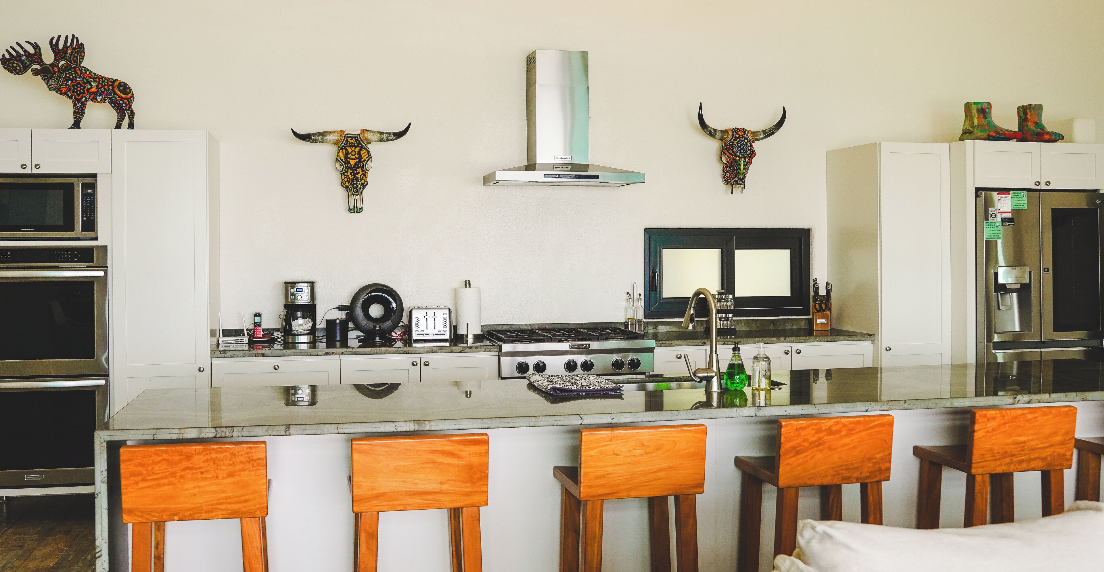 Una cocina moderna con una isla central, taburetes de bar, estufa, horno, microondas y refrigerador. Decoran la pared sobre la estufa cráneos de animales.