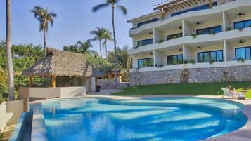 Una piscina con borde curvo, una estructura con techo de paja y un edificio de varios pisos con balcones y plantas de exterior.