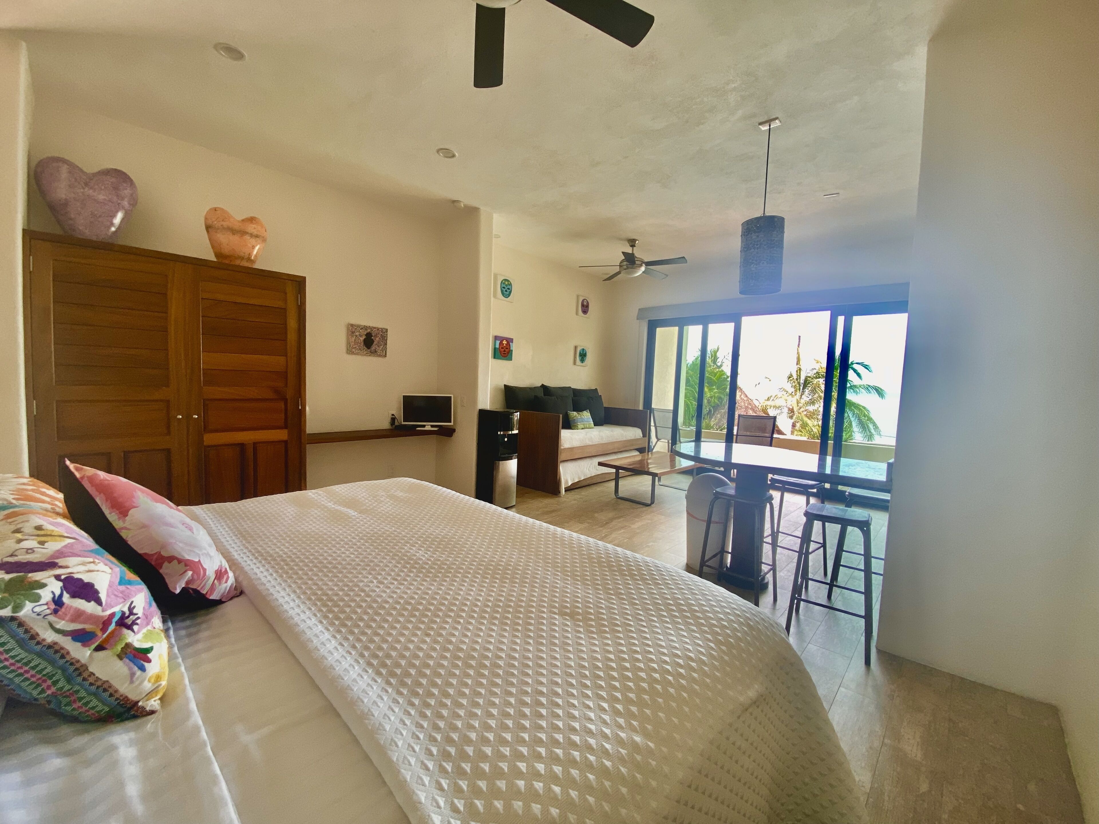 A bedroom with a bed, a desk, a chair, a TV, and a balcony with a view of palm trees.