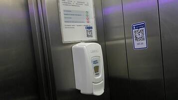 Interior metálico de un ascensor con un código QR, dispensador de desinfectante y un cartel con pautas sobre COVID-19.