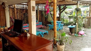A rustic outdoor seating area with wooden tables and benches, a laptop on a desk, and a bamboo structure overhead. - Oasis Hostal Restaurante (Maicao)