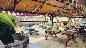 An outdoor patio area with wicker furniture and picnic tables, surrounded by stone flooring and under a wooden pergola with hanging light bulbs. - The Walkhampton Inn (Yelverton)
