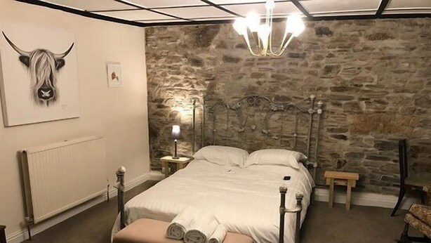 Un dormitorio con pared de piedra, una cama con ropa blanca, un banco, una mesita de noche, una lámpara y una araña.