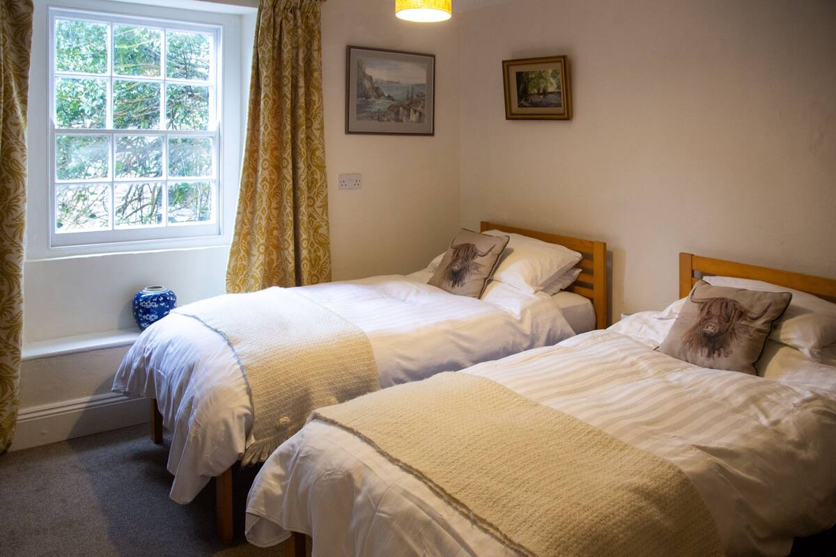 Una habitación con dos camas, una ventana con cortinas, un jarrón azul y dos cuadros en la pared.