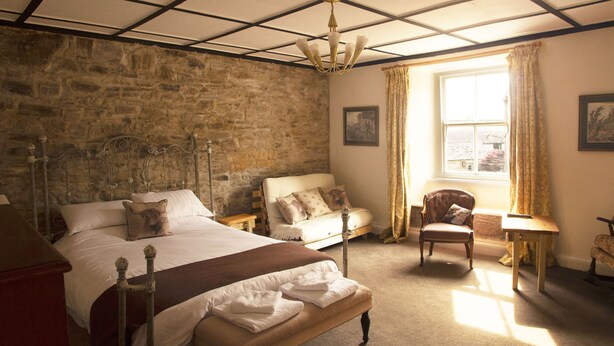 A bedroom with a stone wall, a bed with a headboard, a sofa, a chair, and a window with curtains. - The Walkhampton Inn (Yelverton)