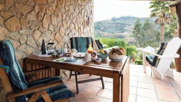A stone wall, a wooden table with breakfast items, a chair, and a view of a hilly landscape.