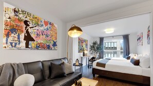 A modern hotel room with a black leather sofa, a bed with white bedding, a coffee table with a book, and a large abstract painting on the wall. -  LiV MTL - Avenue (Montreal)