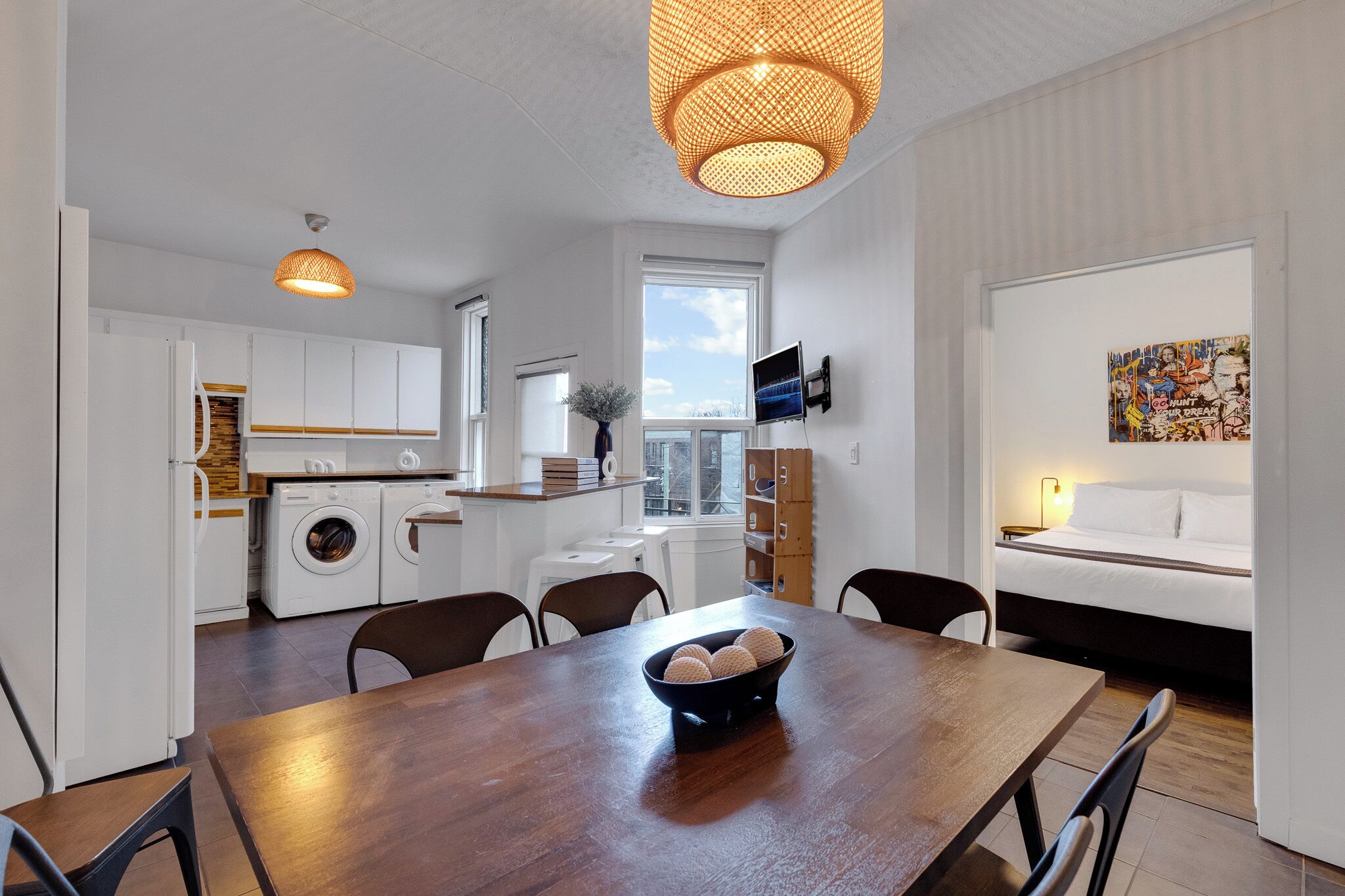 Una cocina moderna con armarios blancos, una mesa de comedor con un tazón de fruta y una cama con un televisor al fondo.