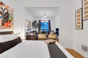 Una habitación de hotel moderna con una cama grande, un área de descanso con dos sillones, una mesita, una planta y una ventana con cortinas.