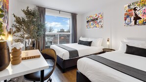 A hotel room with two beds, a desk, a chair, a potted plant, and artwork on the walls. -  LiV MTL - Avenue (Montreal)