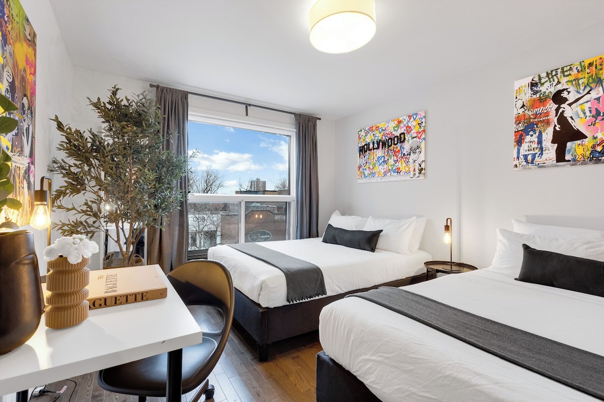 A hotel room with two beds, a desk, a chair, a potted plant, and artwork on the walls.