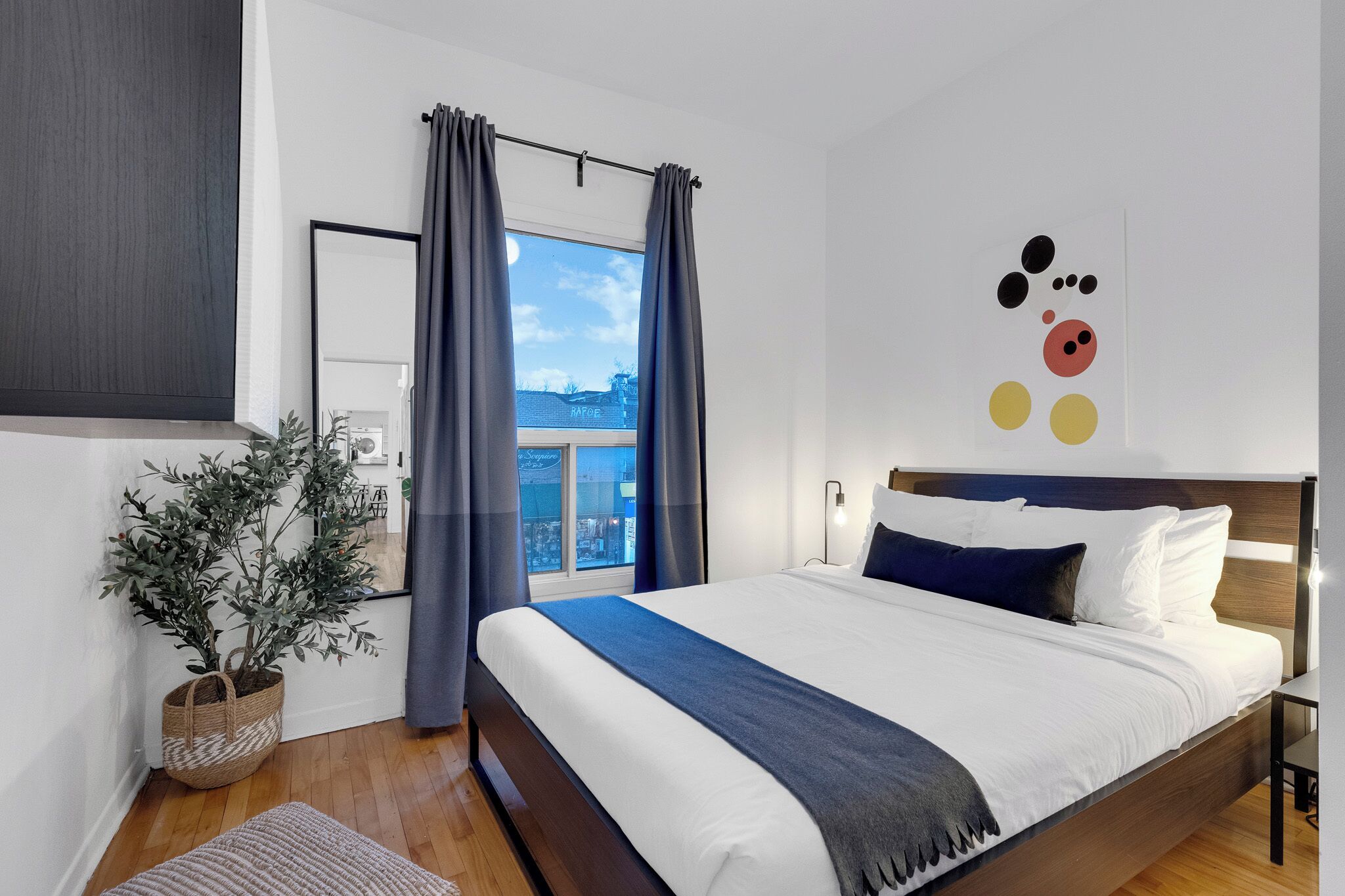 A modern bedroom with a large bed, a potted plant, a view through a window, and a wall art piece.