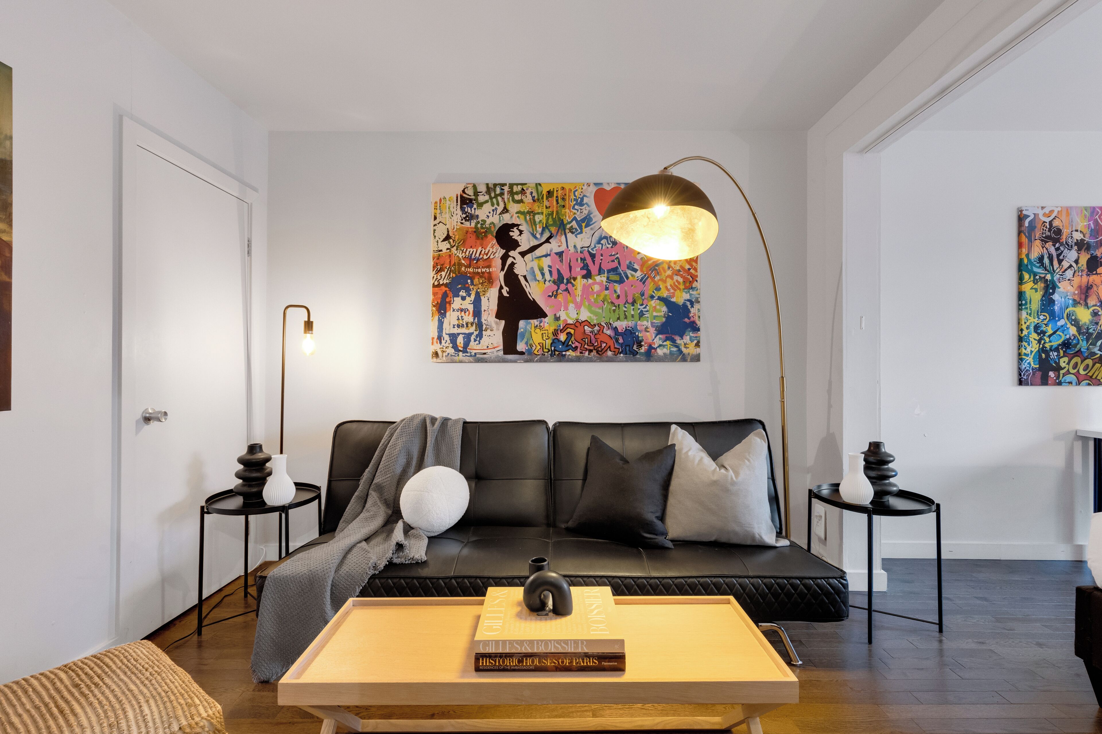 A modern living room with a black leather sofa, a coffee table with books, a floor lamp, and a colorful abstract painting on the wall.