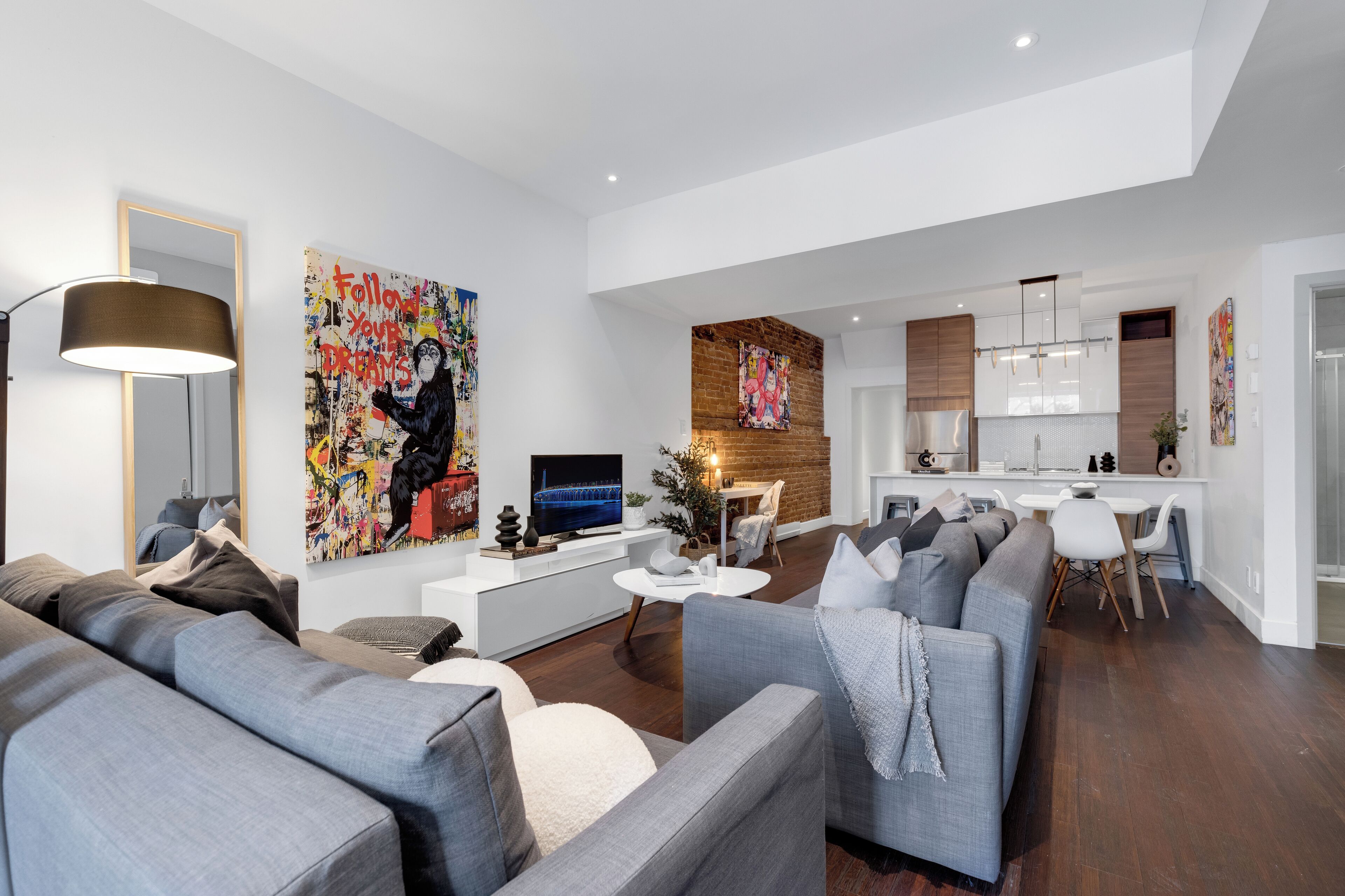 Un salon moderne avec un canapé gris, une table basse et un téléviseur. On y trouve également un coin cuisine avec un mur en briques et des meubles blancs.