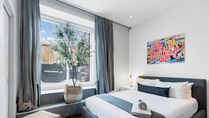 Camera da letto moderna con un letto grande, una finestra con tende, una pianta e un'opera d'arte colorata al muro.