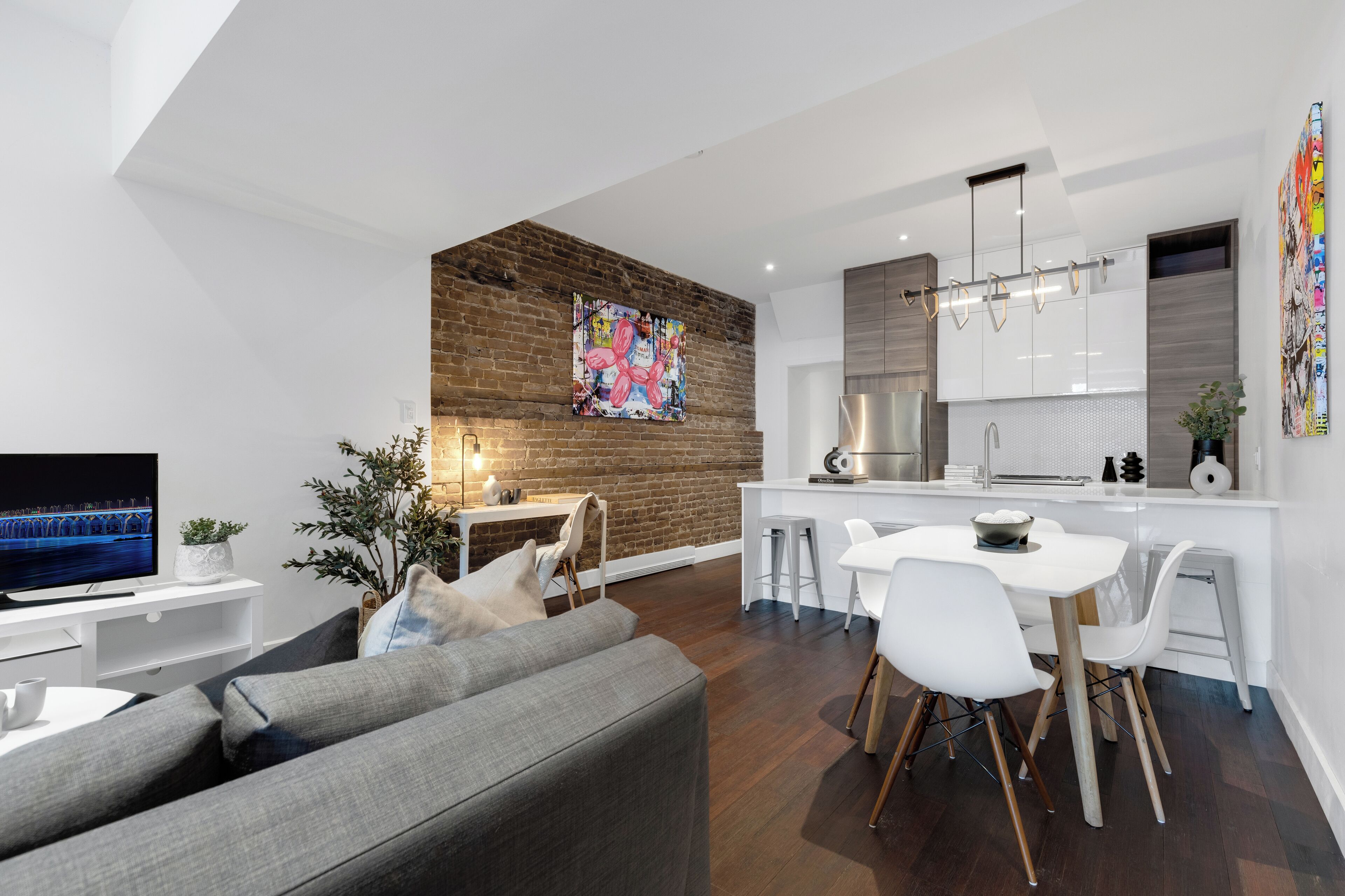 A modern living room with a grey sofa, a TV, and a dining area with white chairs and a table.