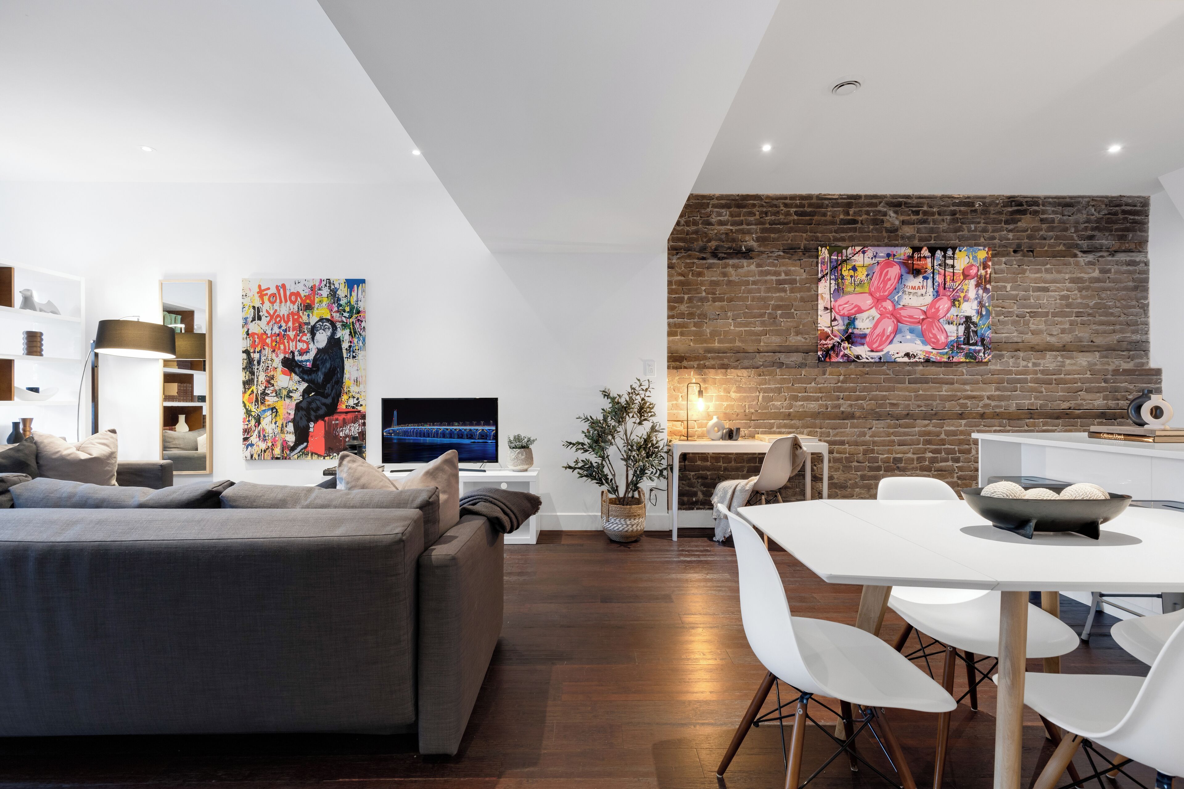 A modern living room with a grey sofa, a dining table with white chairs, a TV, and two large abstract paintings on the brick wall.