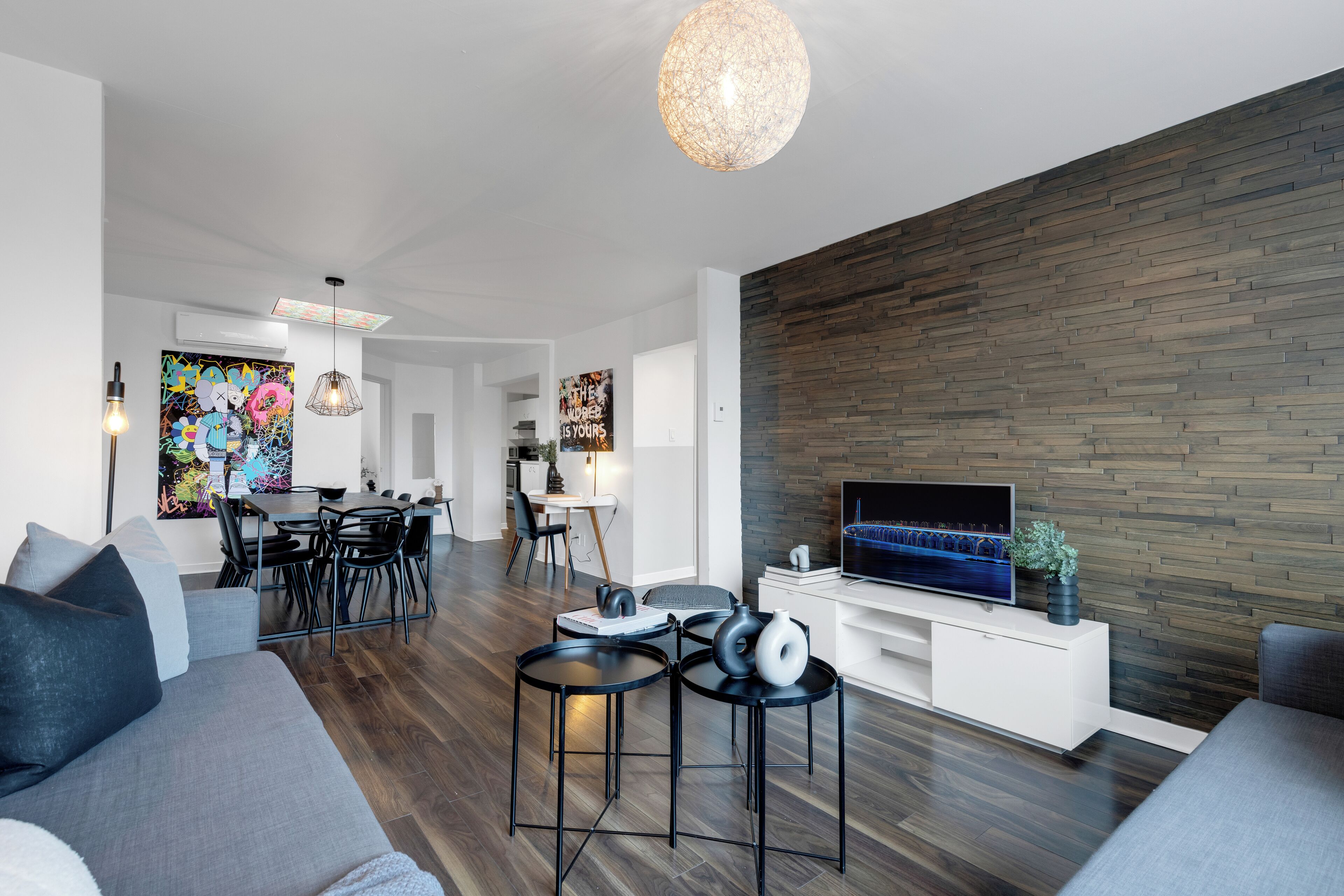 Uma sala moderna com televisão de tela plana, área de jantar com mesa e cadeiras, e cozinha ao fundo.