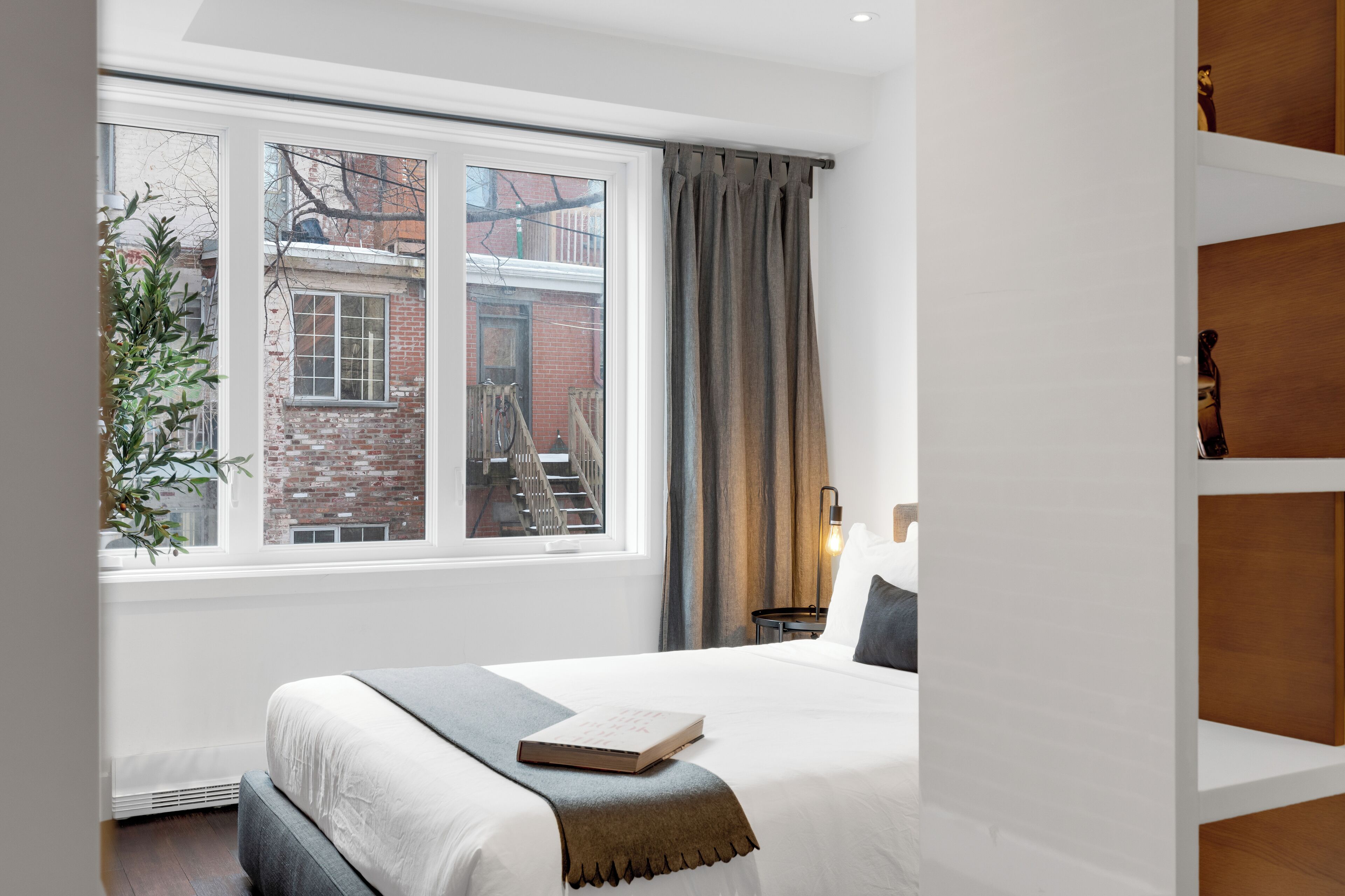 A bedroom with a large window, a bed, a bedside table, and a nightstand.