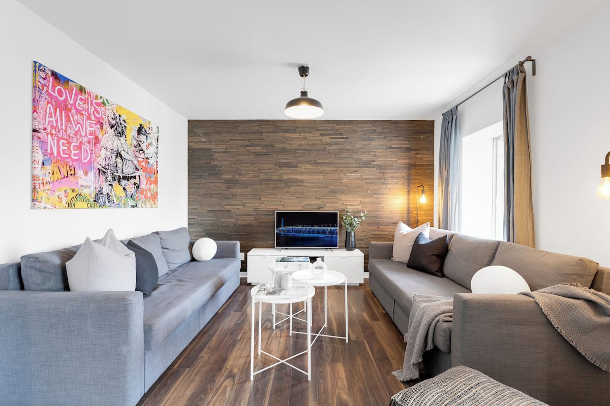 A modern living room with two grey sofas, a white coffee table, a flat-screen TV on a wooden wall, and a large colorful artwork.